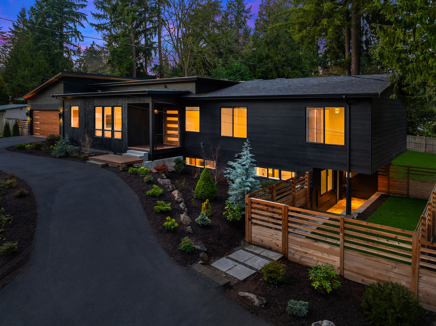Modern two-story house with large windows glowing warmly at dusk, surrounded by trees and landscaped garden, with a curved driveway and wooden fence in the foreground.