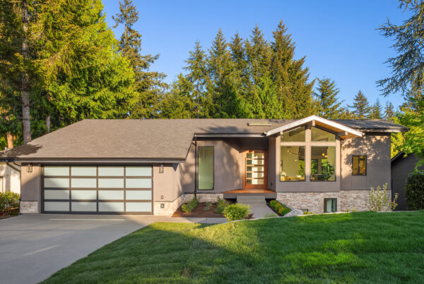 Modern single-story house with large front windows, a two-car garage with frosted glass doors, a stone accent wall, and a well-kept lawn, surrounded by tall trees under a clear blue sky.