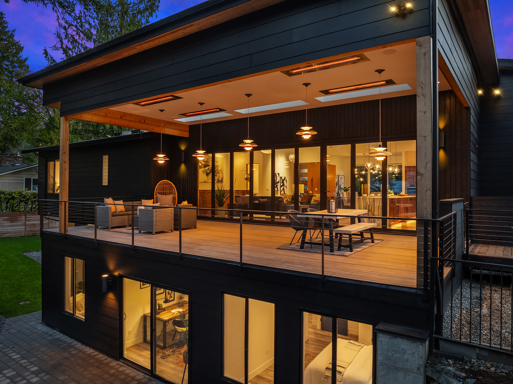 A modern two-story house at dusk with large glass doors, a spacious covered balcony with warm hanging lights, outdoor seating, and visible interior living and bedroom areas through illuminated windows.