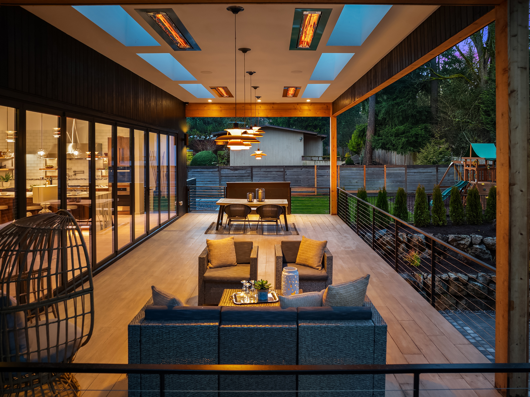 Modern covered patio with skylights, stylish outdoor seating, dining area, hanging chair, and warm lighting; large windows show a kitchen inside. Backyard and playset visible in the background.