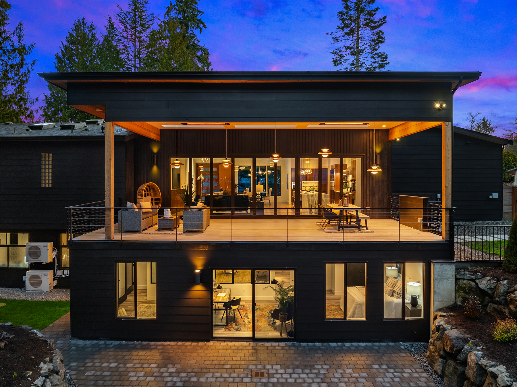 Modern two-story house at dusk with large windows, a spacious upper deck featuring seating and dining areas, warm indoor lighting, and surrounding trees and stone landscaping.