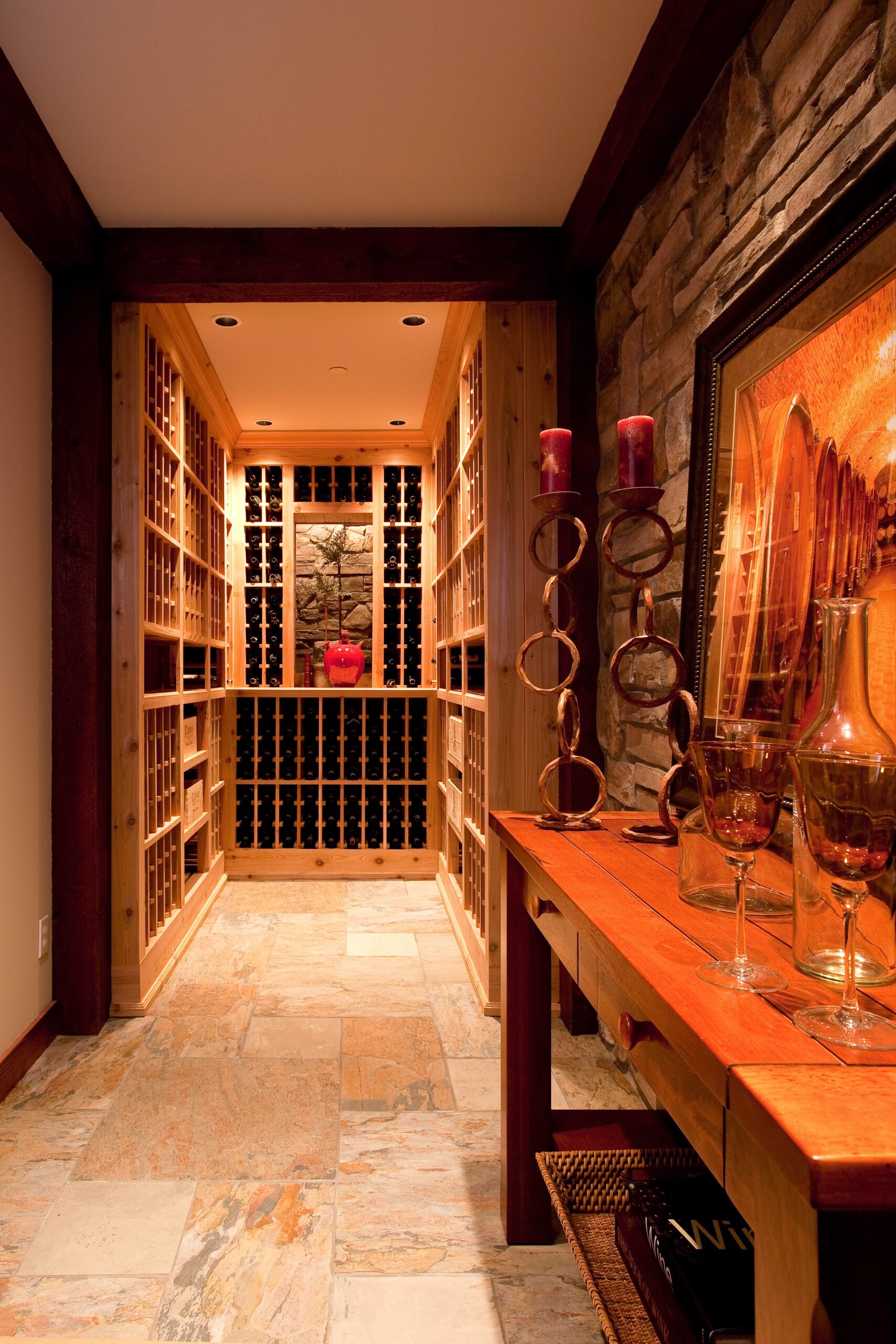 A warmly lit wine cellar with wooden racks filled with wine bottles, stone tile flooring, and a wooden table displaying glassware, decorative candles, and framed artwork.
