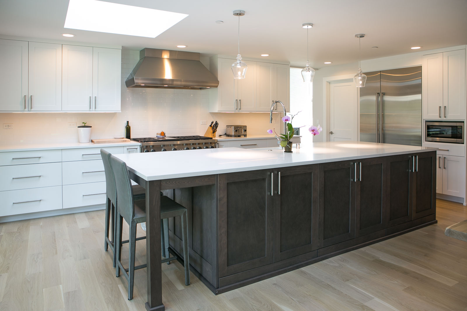 Womack-10 Modern kitchen with white cabinets, stainless steel appliances, a large dark wood island with white countertop, two bar stools, pendant lights, and light wood flooring. A potted plant and orchid decorate the island.