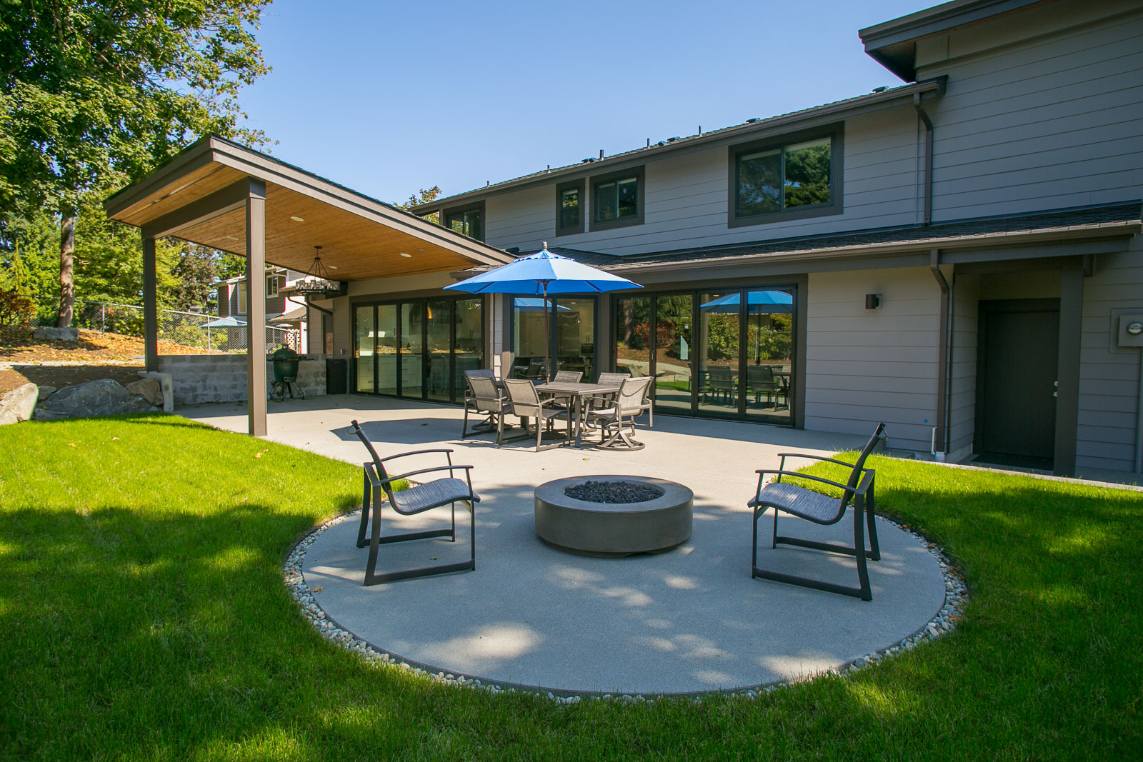 Womack-3 Modern backyard patio with round fire pit, four cushioned chairs, and a shaded dining area with a blue umbrella. The patio is surrounded by green grass and attached to a contemporary gray house.