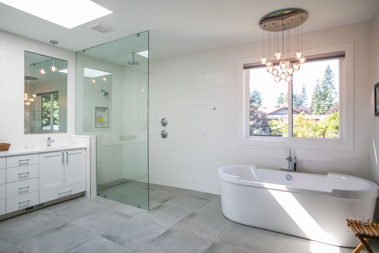 Womack-4 Modern bathroom with a glass-enclosed shower, white freestanding bathtub under a window, white vanity with sink on the left, gray tiled floor, and a contemporary chandelier on the ceiling.