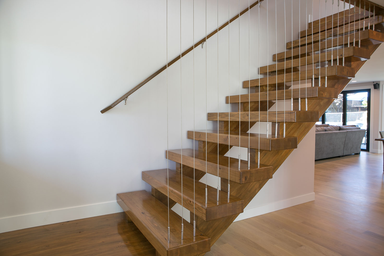Womack-6 A modern wooden staircase with open risers and thin vertical metal railings is shown against a white wall in a bright, contemporary home interior with hardwood floors.