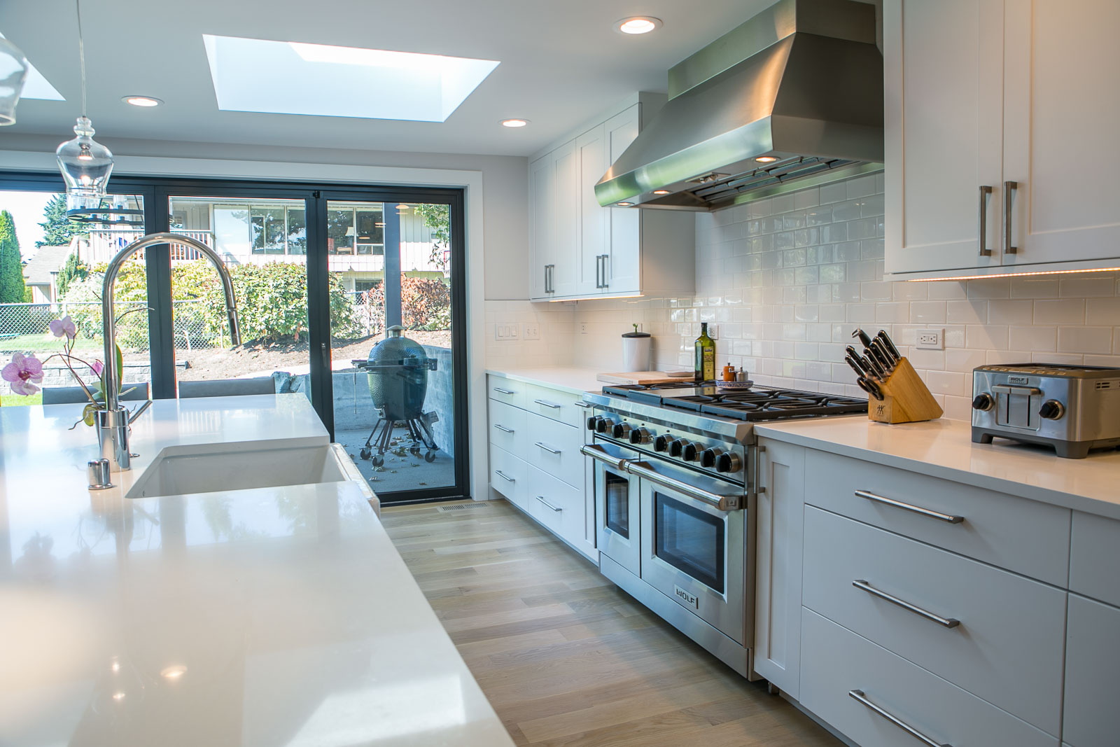 Womack-8 Modern kitchen with white cabinets, stainless steel appliances, a large gas stove, knife block, toaster, and sliding glass doors leading to a patio with a barbecue grill outside. Light wood flooring and bright natural light.