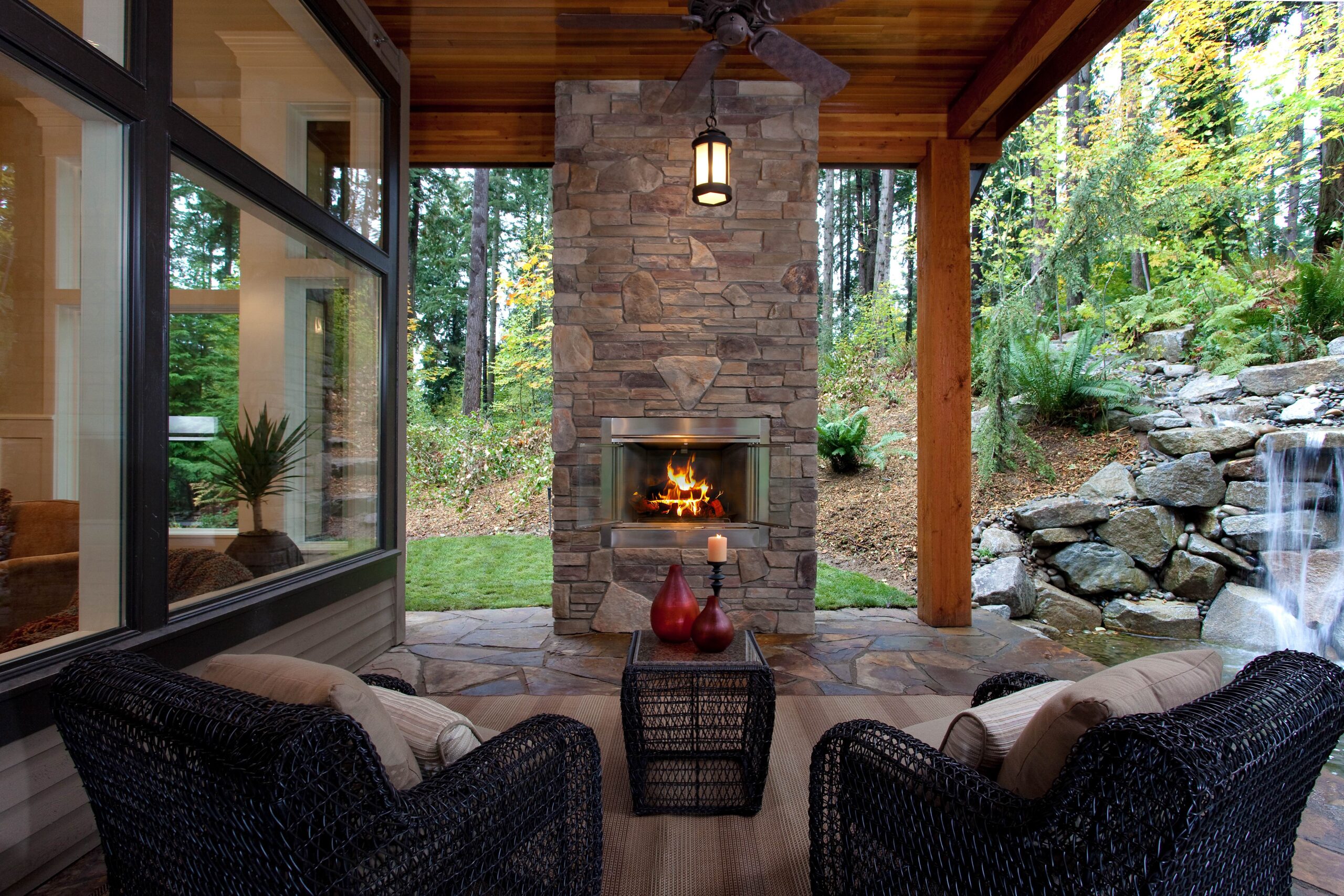 Covered outdoor patio with two wicker chairs facing a stone fireplace with a lit fire. Large windows and ceiling fan overhead, surrounded by lush green trees and a waterfall feature to the right.