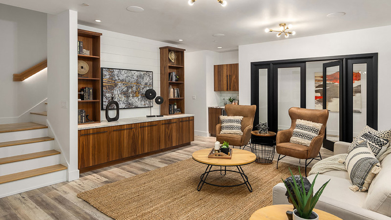 Modern living room with wood accents, a large woven rug, cozy armchairs, a white sofa, built-in shelves, wall art, and a TV. Stairs lead up on the left, and black-framed glass doors open to another room.